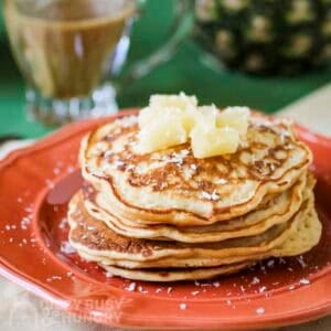 Side shot of a stack of pineapple pancakes garnished with coconut shreds and pineapple chunks on an orange plate.
