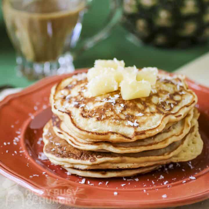 Side shot of a stack of pineapple pancakes garnished with coconut shreds and pineapple chunks on an orange plate.