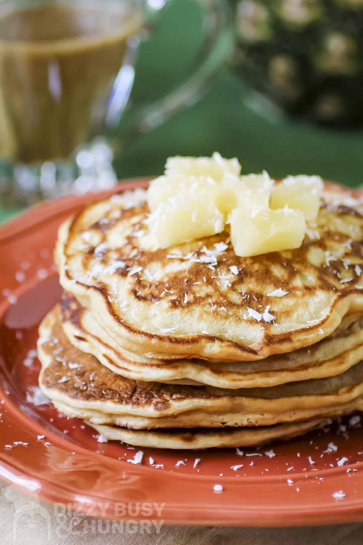Side shot of a stack of pineapple pancakes garnished with coconut shreds and pineapple chunks on an orange plate.