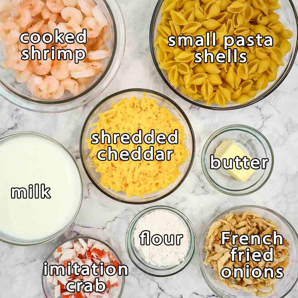 Overhead shot of ingredients - cooked shrimp, small pasta shells, shredded cheddar, milk, butter, flour, french fried onions, and imitation crab.