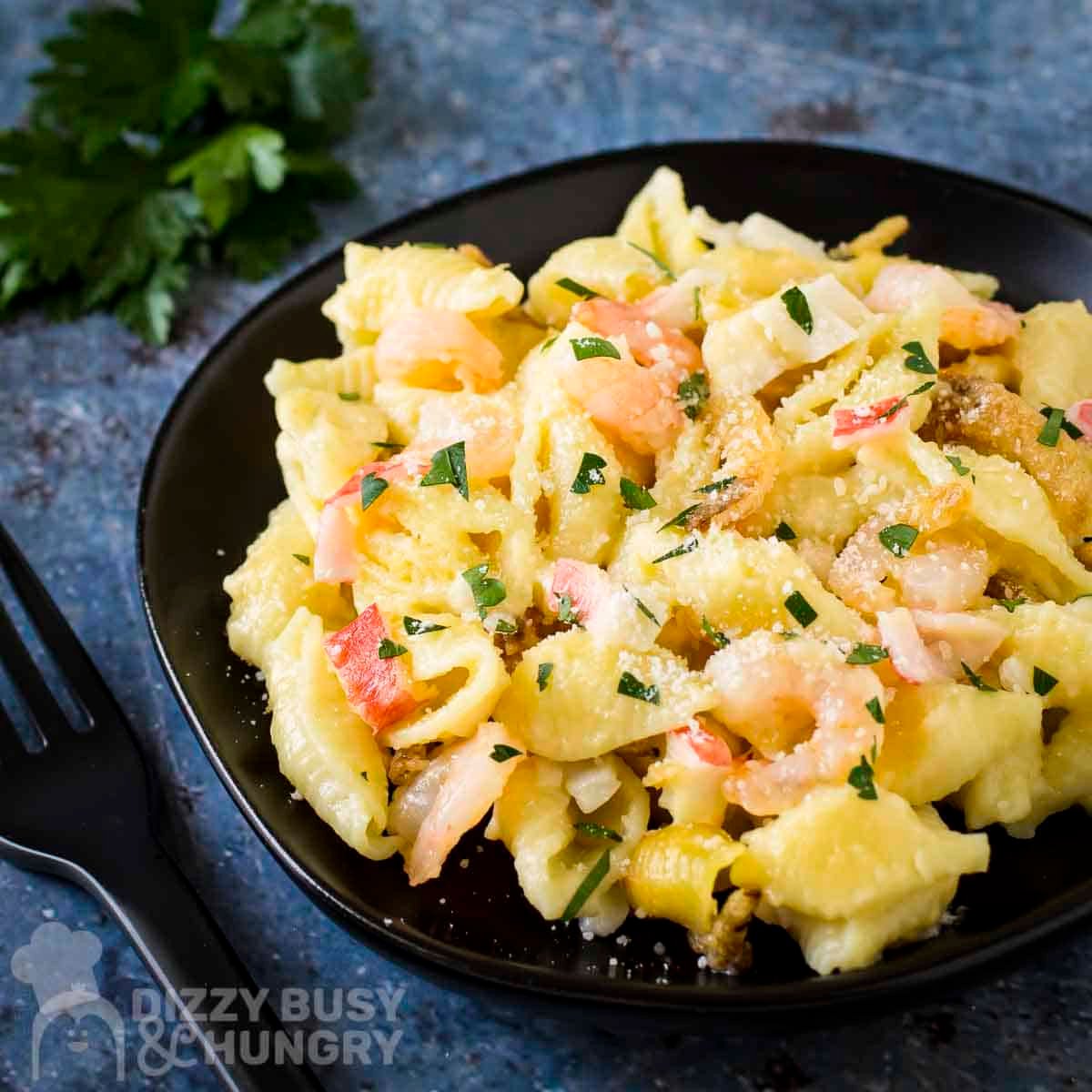 Side shot of seafood mac and cheese garnished with fresh herbs on a black plate with more fresh herbs in the background.