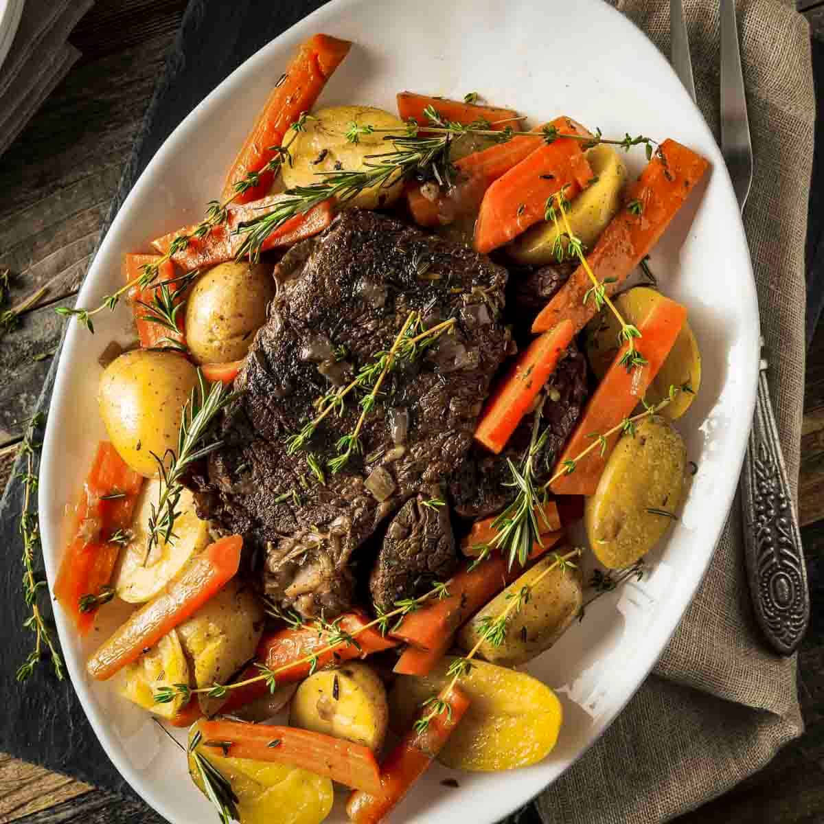 Overhead shot of braised chuck roast surrounded by carrots and potatoes on a white serving plate garnished with fresh rosemary.
