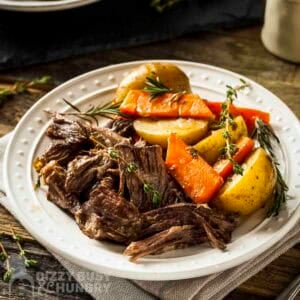 Side shot of braised pot roast with carrots and potatoes on a white plate garnished with fresh rosemary.