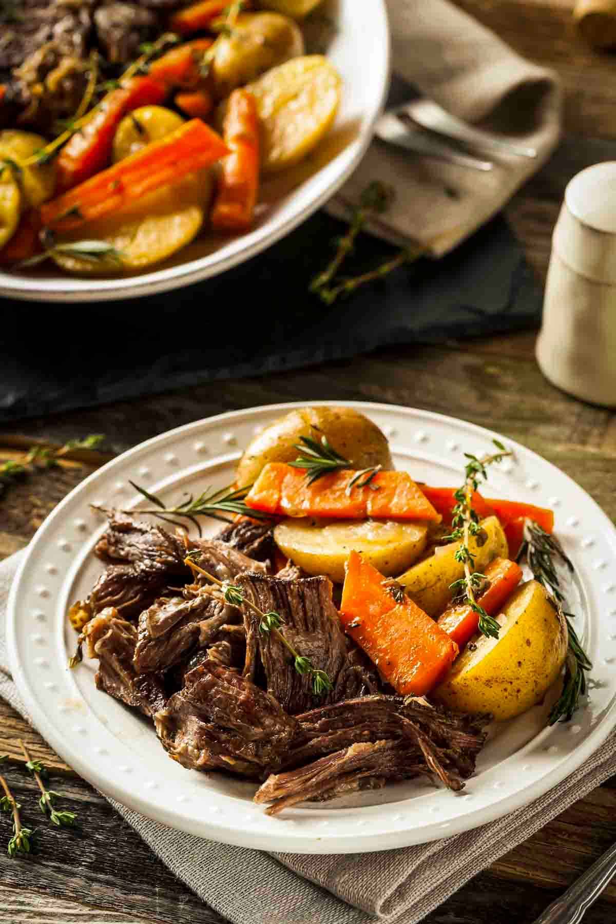 Side shot of braised pot roast with carrots and potatoes on a white plate garnished with fresh rosemary.