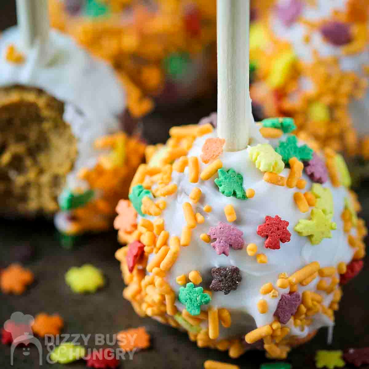 Side close up shot of a pumpkin cake pop with fall spinkles on a black surface with more cake pops in the background.
