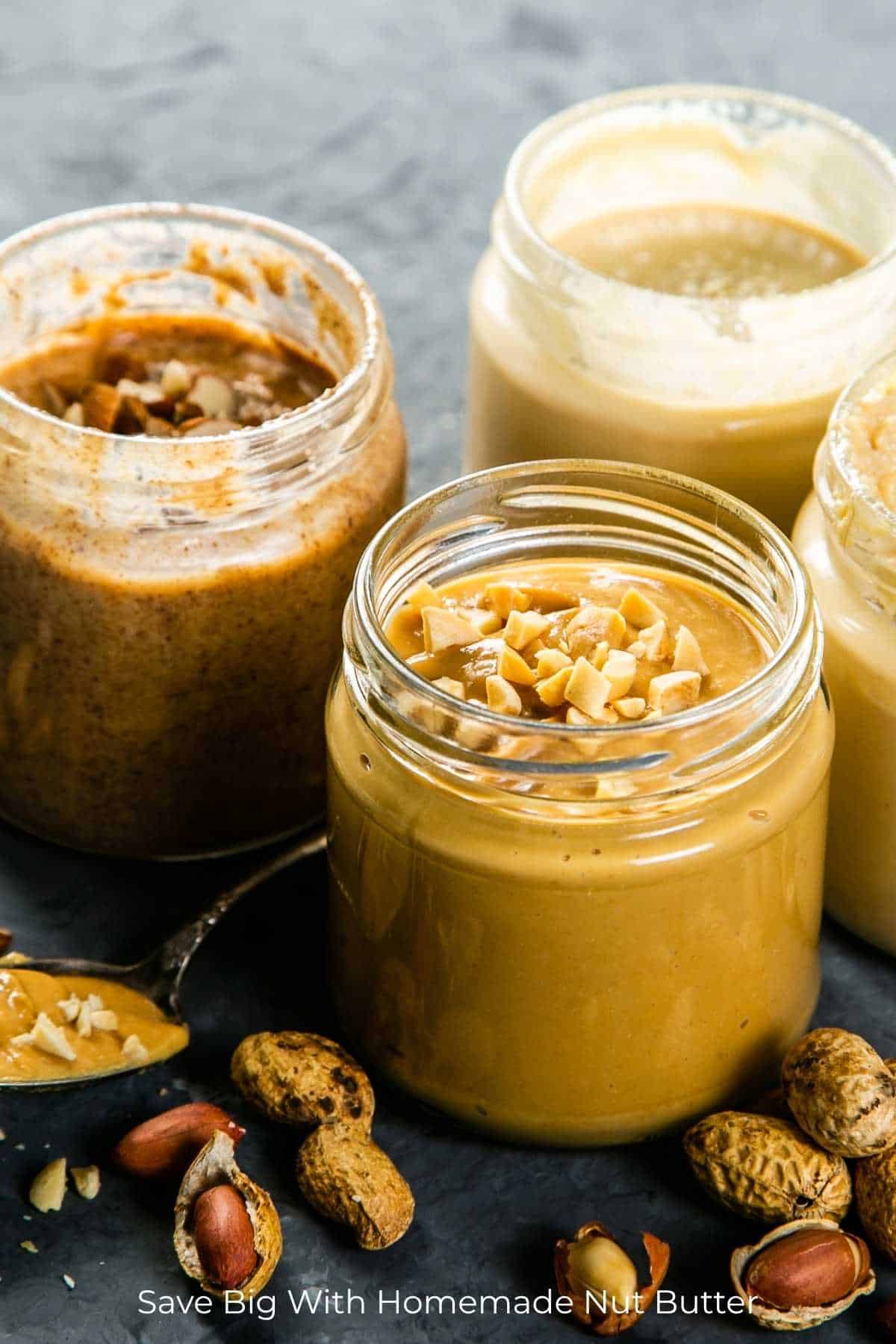 Four creamy homemade nut butters with chopped nut toppings on a dark counter with various unprocessed nuts strewn over the counter.