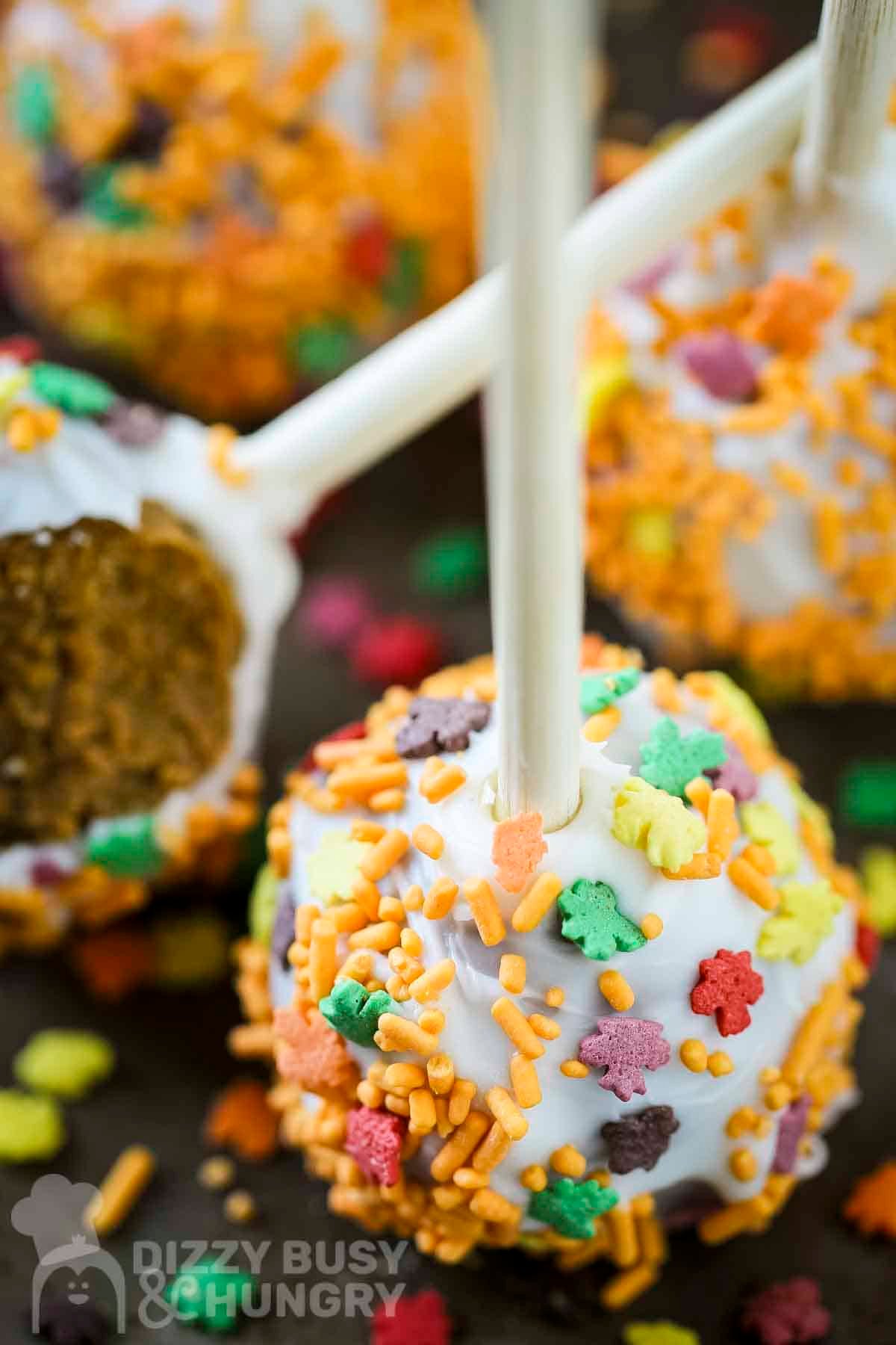 Side close up shot of a pumpkin cake pop with fall spinkles on a black surface with more cake pops in the background.