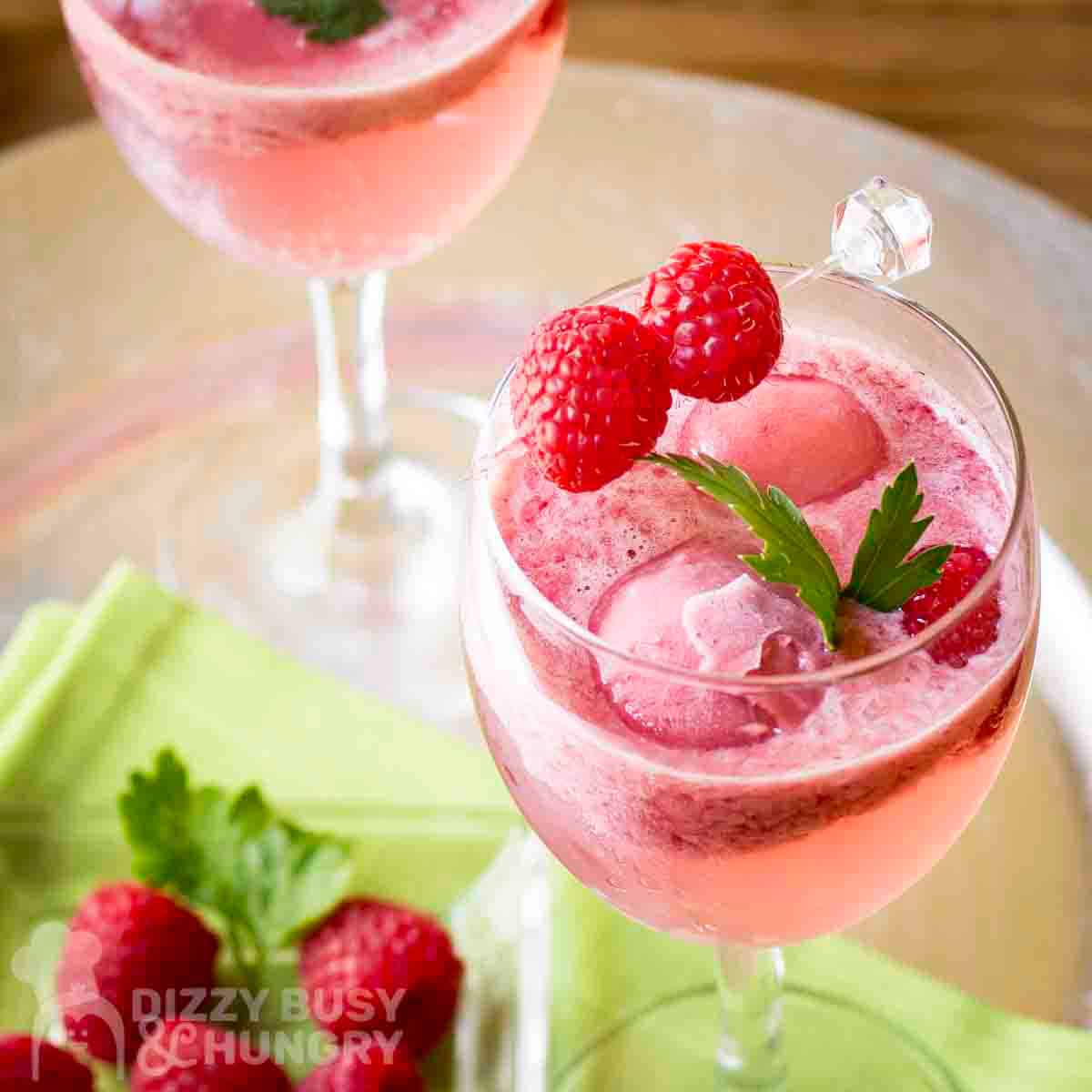Close up shot of two cocktail glasses with raspbery spritz garnished with fresh mint and raspberries with more raspberries and a green cloth underneath.