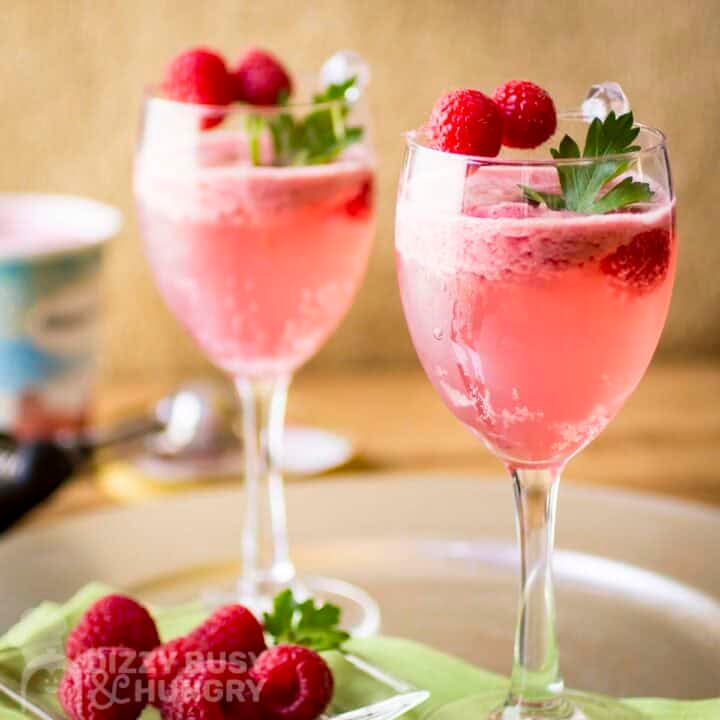Side shot of two cocktail glasses with raspbery spritz garnished with fresh mint and raspberries with more raspberries and a green cloth underneath.