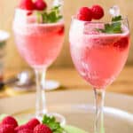 Side shot of two cocktail glasses with raspbery spritz garnished with fresh mint and raspberries with more raspberries and a green cloth underneath.