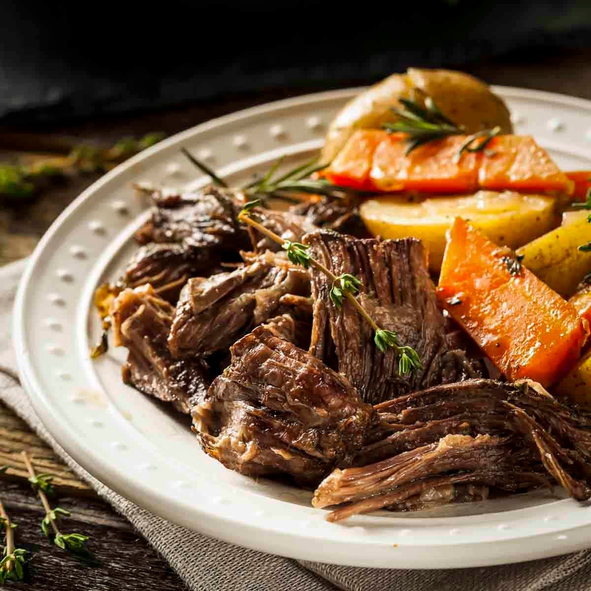 Side shot of braised pot roast with carrots and potatoes on a white plate garnished with fresh rosemary.