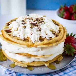 Side shot of waffle ice cream cake garnished with whipped cream and chopped nuts on a grey plate with strawberries on the side.