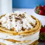 Side shot of waffle ice cream cake garnished with whipped cream and chopped nuts on a grey plate with strawberries on the side.