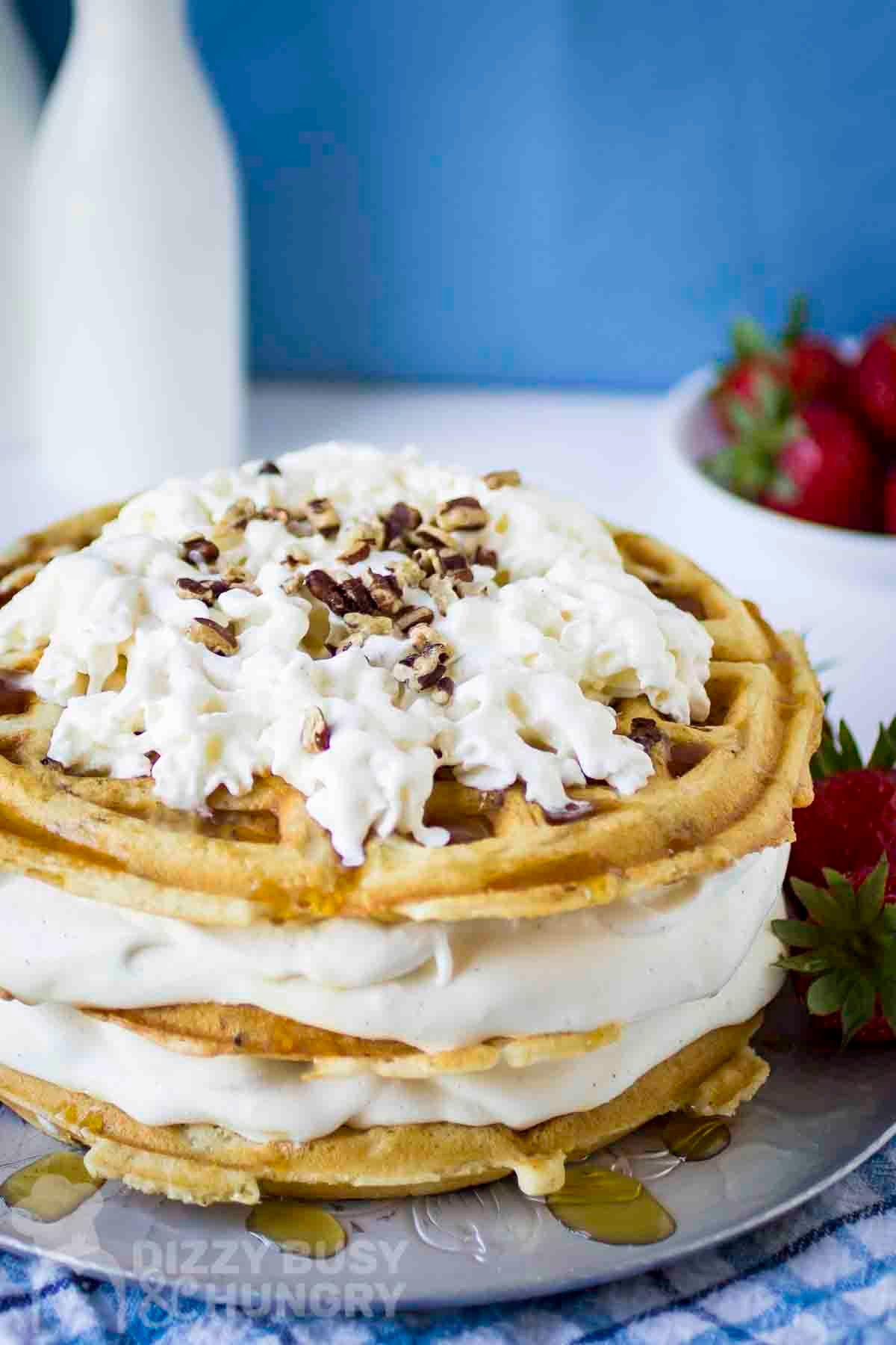 Side shot of waffle ice cream cake garnished with whipped cream and chopped nuts on a grey plate with strawberries on the side.
