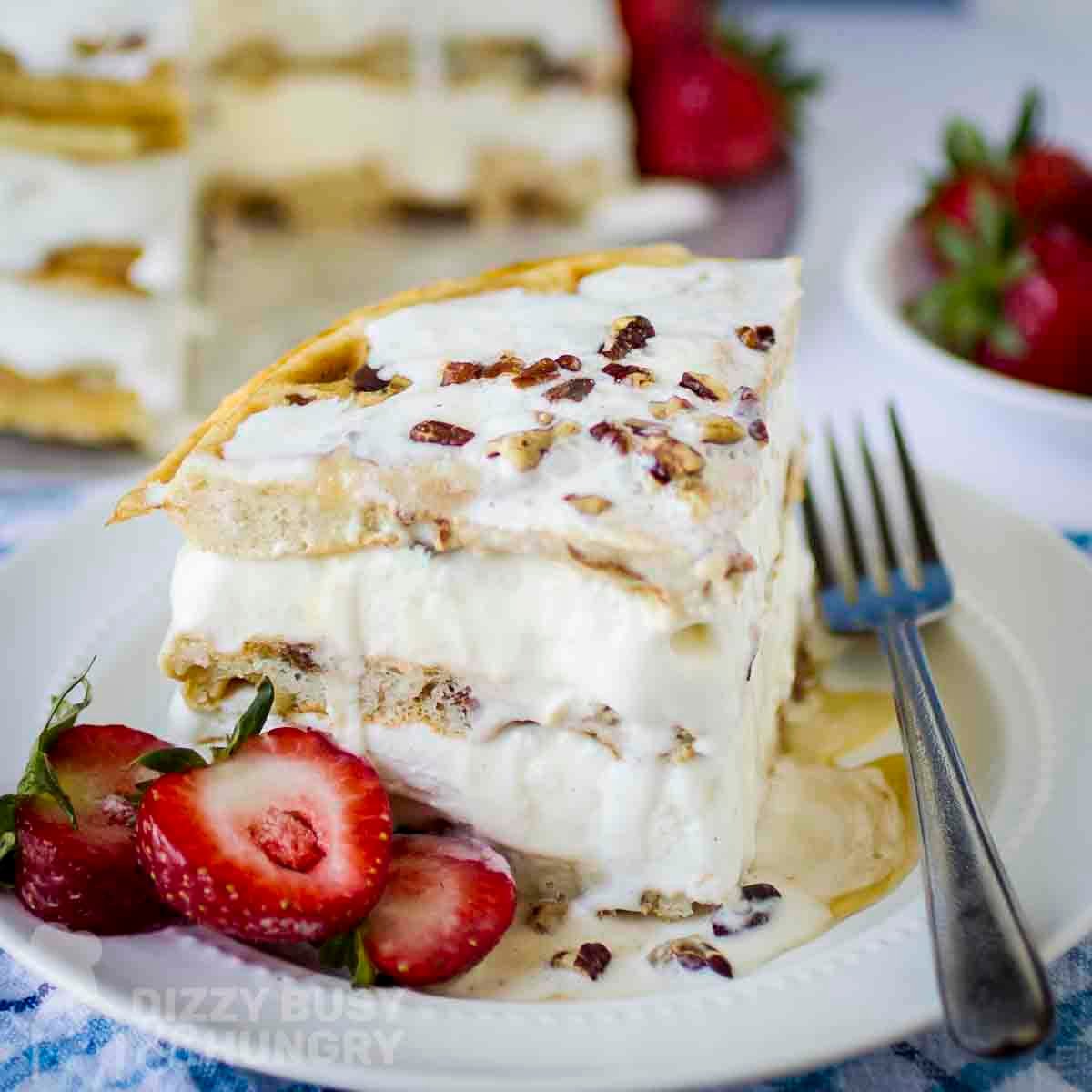 Side shot of a quarter of a waffle ice cream cake garnished with whipped cream and chopped nuts on a white plate with strawberries on the side.