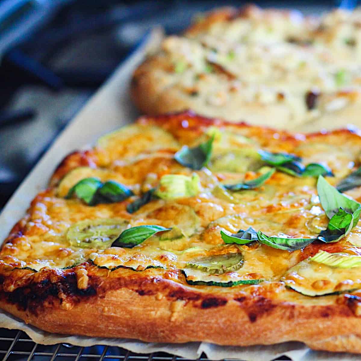 Side shot of Zucchini Flower Pizza  with cheese, zucchini, and basil toppings on a piece of parchment paper on a blue tile surface.