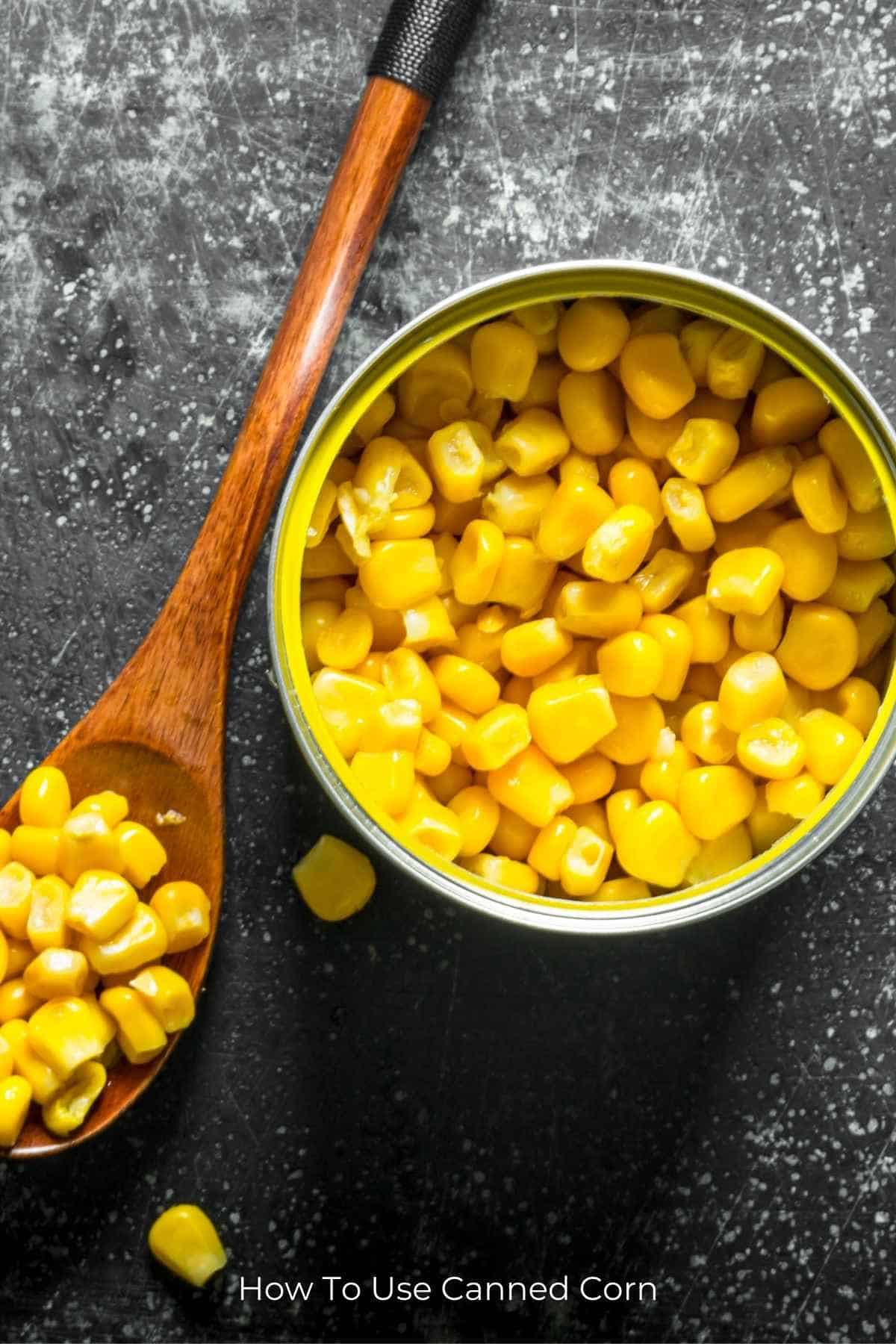 Overhead view of an open can of corn with a wooden spoon to the right holding a bunch of kernals all on a charcoal gray background.