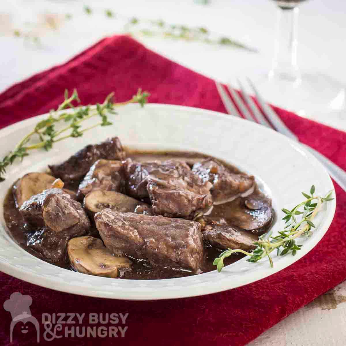 Side shot of beef stew in a white bowl with fresh thyme on the sides all on a red cloth.