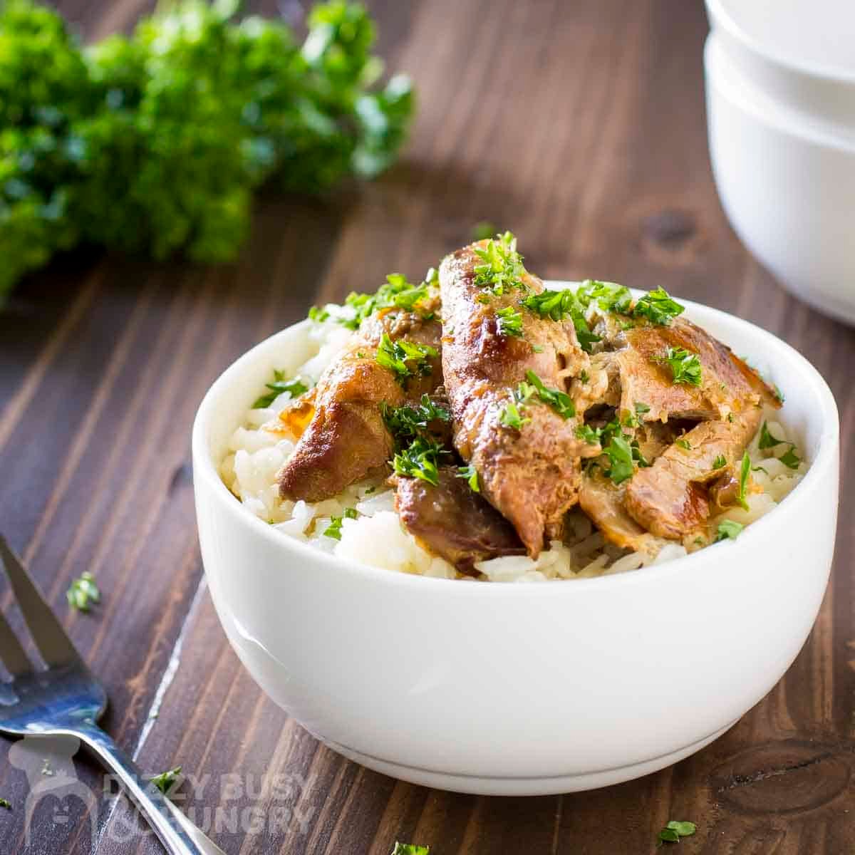 Side shot of beer chicken on a bed of rice in a white bowl garnished with fresh herbs with a fork and more herb sprigs in the background.