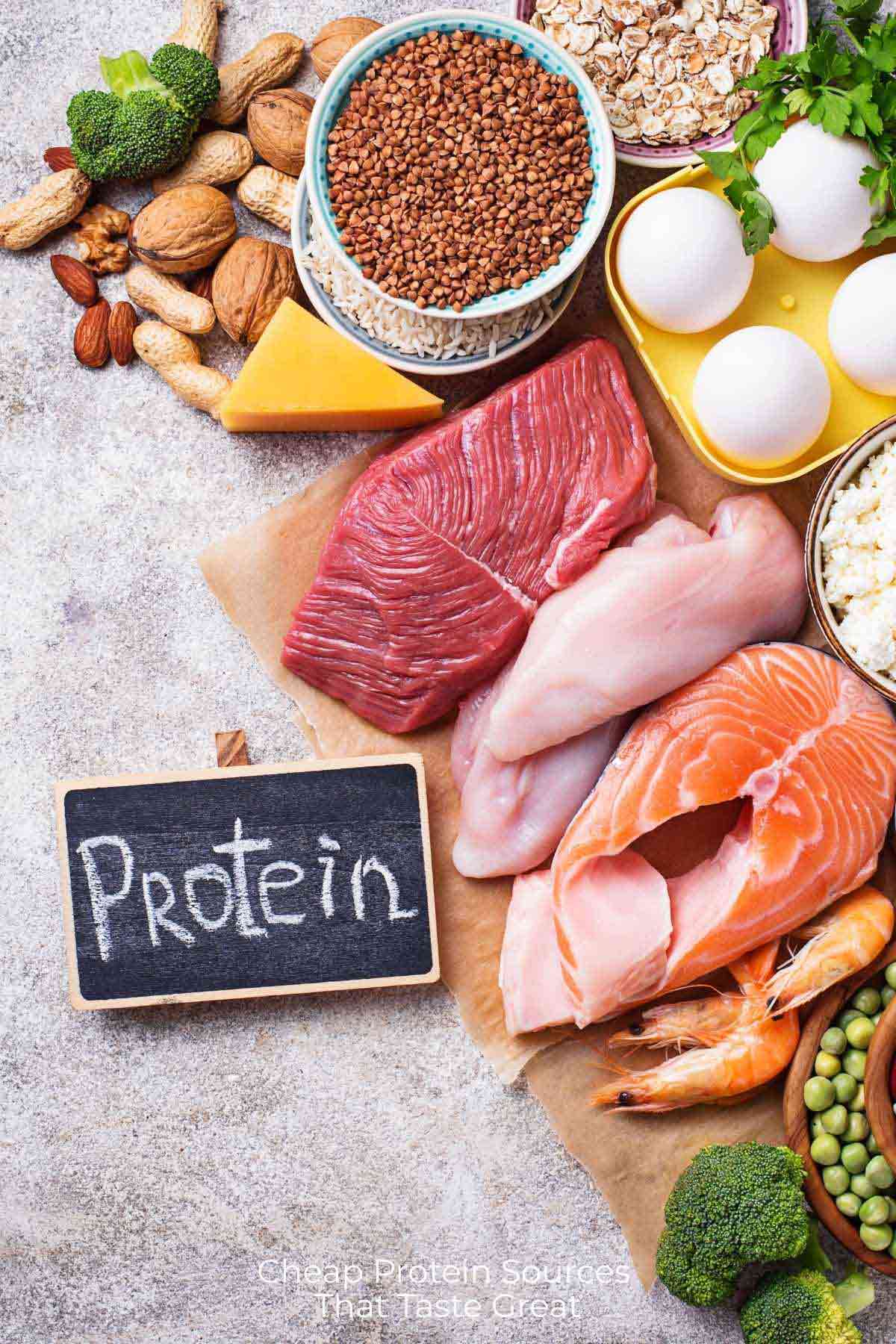 Overhead view of possible cheap protein sources like eggs and lentils on a gray concrete counter with a small chalkboard to the side that has the word 'protein' written on it.