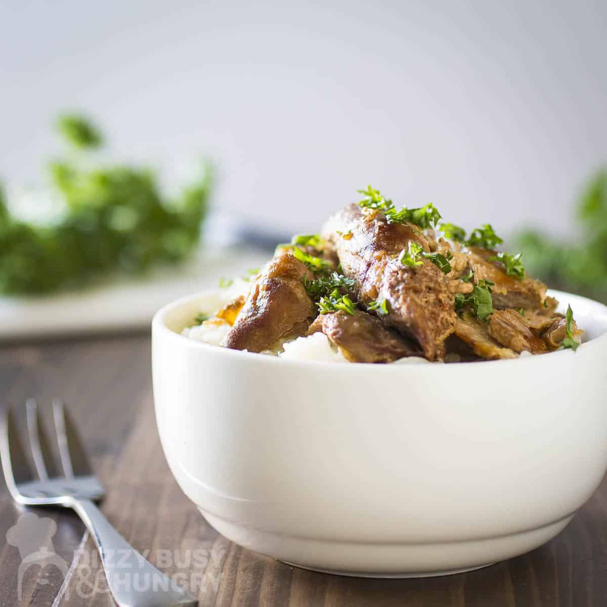 Side shot of beer chicken on a bed of rice in a white bowl garnished with fresh herbs with a fork and more herb sprigs in the background.