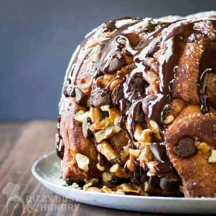 Side shot of whole monkey bread sprinkled with powdered sugar and chocolate syrup on a white plate on a wooden table.