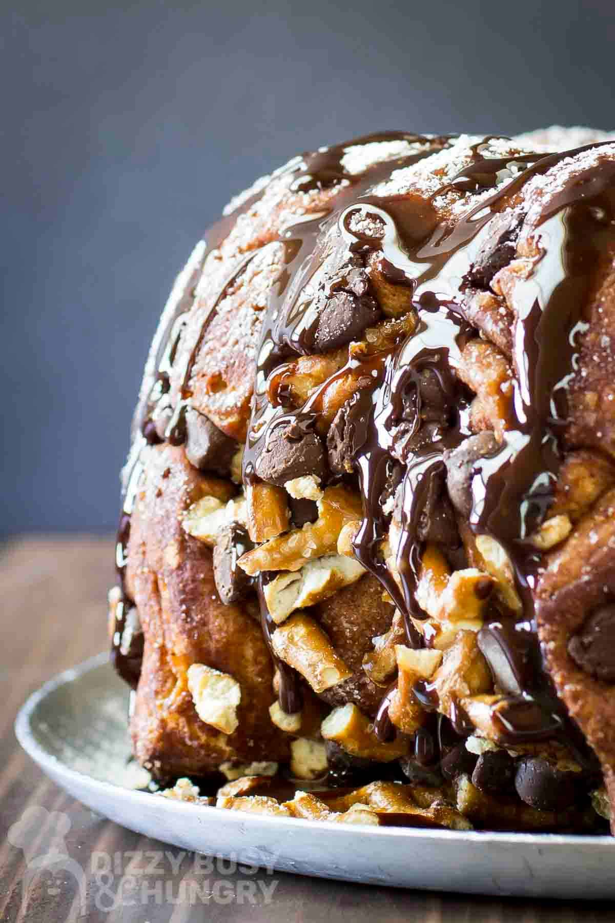 Side shot of whole monkey bread sprinkled with powdered sugar and chocolate syrup on a white plate on a wooden table.