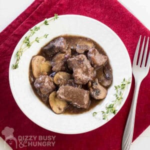 Overhead shot of beef stew in a white bowl with fresh thyme on the sides all on a red cloth with a fork to the right.
