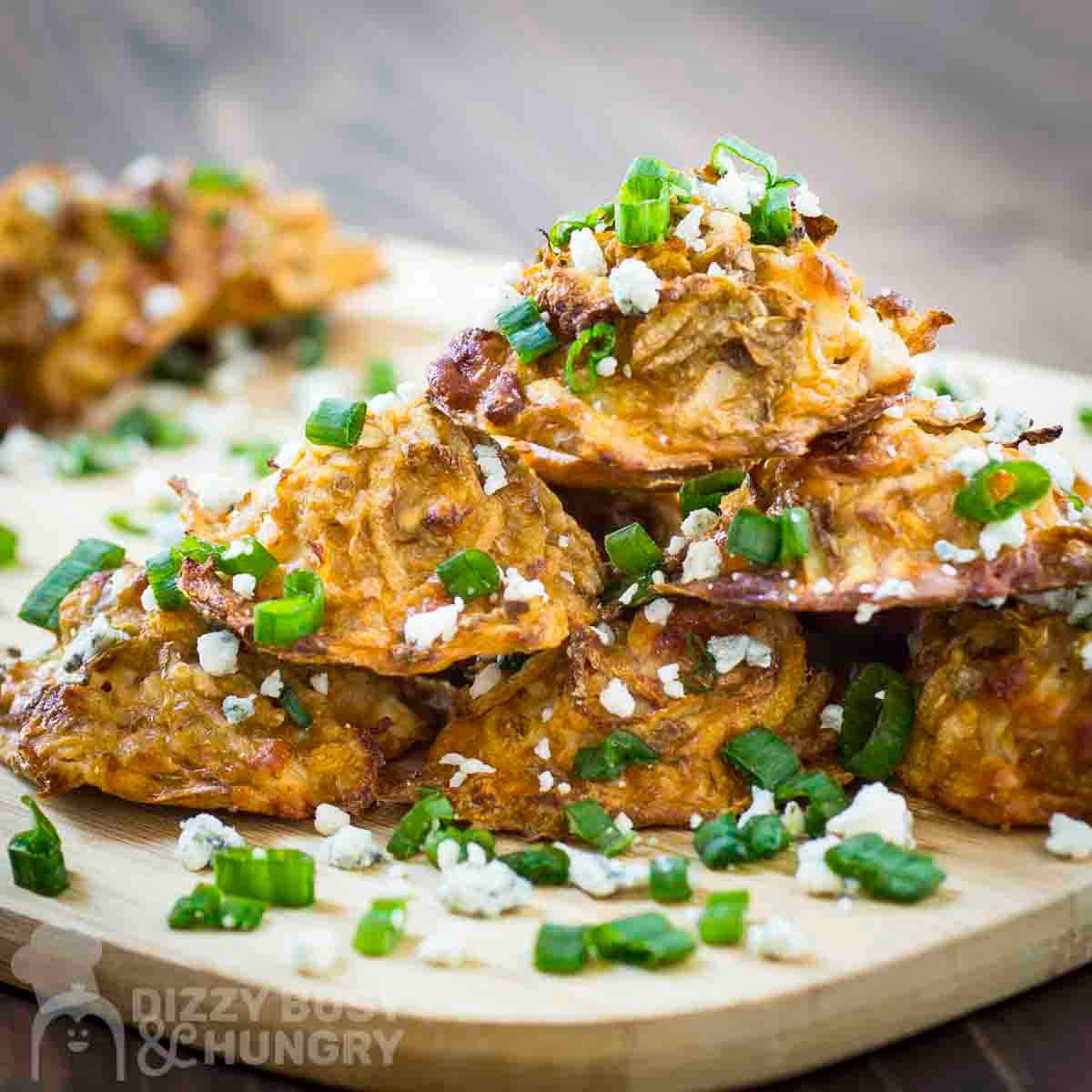 Side shot of multiple potato bites stacked on a wooden cutting board garnished with blue cheese crumbles and green onions.