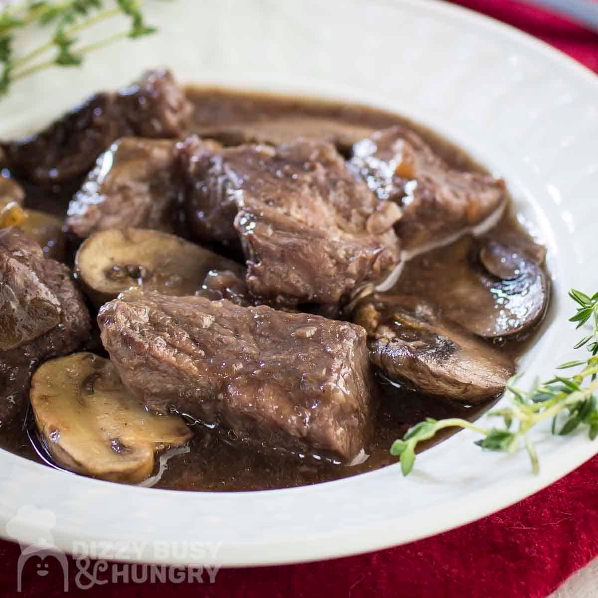 Side shot of beef stew in a white bowl with fresh thyme on the sides all on a red cloth.