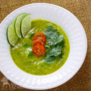 Overhead shot of zucchini curry soup garnished with tomatoes, parsley, and lime slices in a white bowl on a white plate.