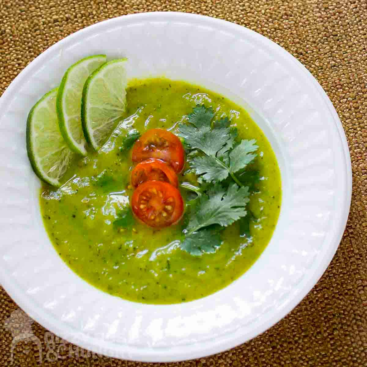 Side shot of zucchini curry soup garnished with tomatoes, parsley, and lime slices in a white bowl.