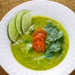 Overhead shot of zucchini curry soup garnished with tomatoes, parsley, and lime slices in a white bowl on a white plate.