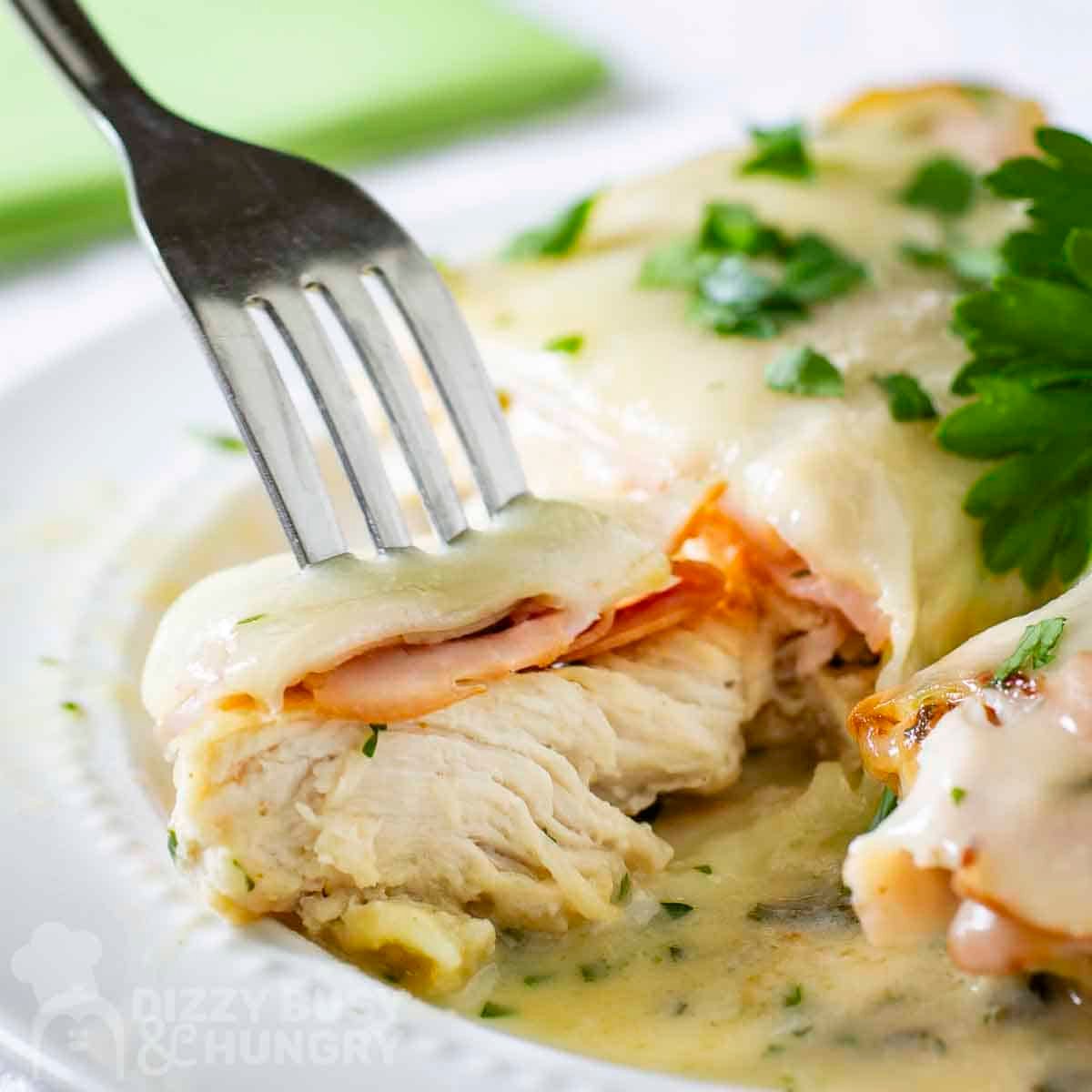 Side close up shot of a fork holding a piece of chicken cordon bleu on a white plate.