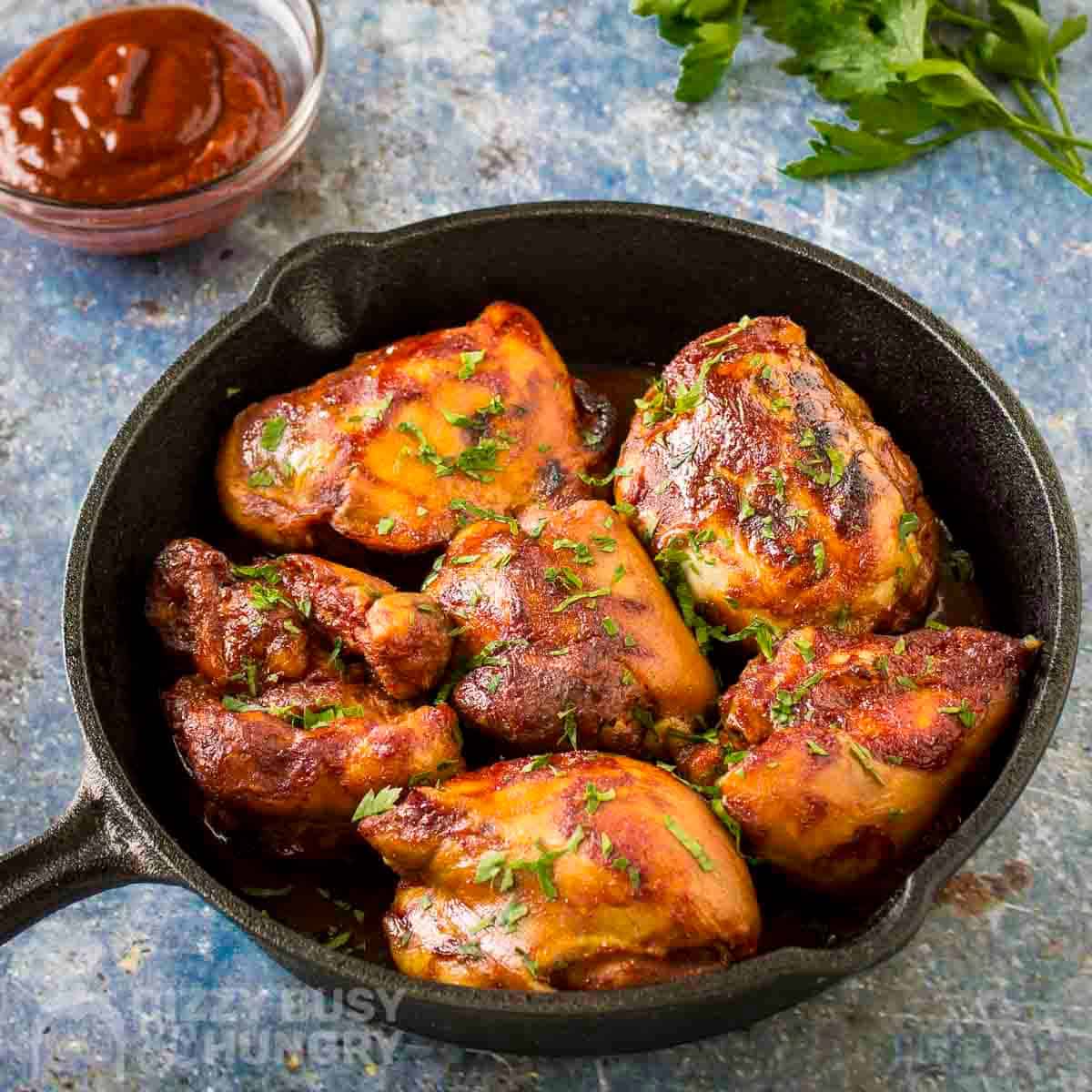 Side angled shot of BBQ chicken thighs in a cast iron skillet garnished with fresh parsley with more fresh parsley sprigs in the background.