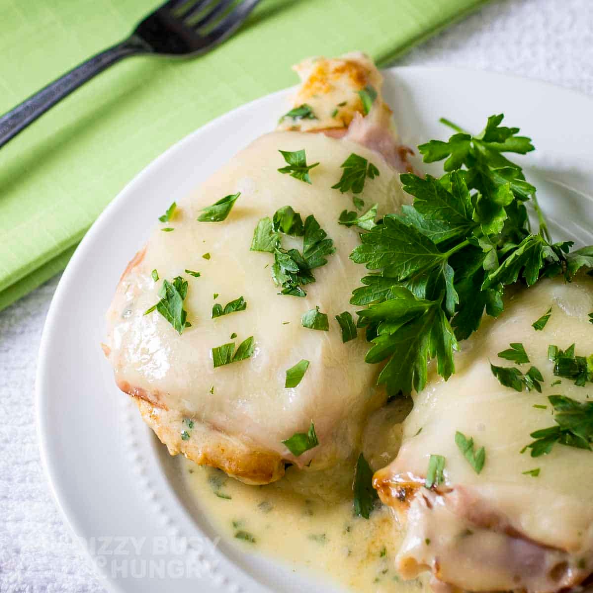 Close up shot of two pieces of chicken cordon bleu garnished with fresh parsley on a white plate with a fork on a green cloth on the side.