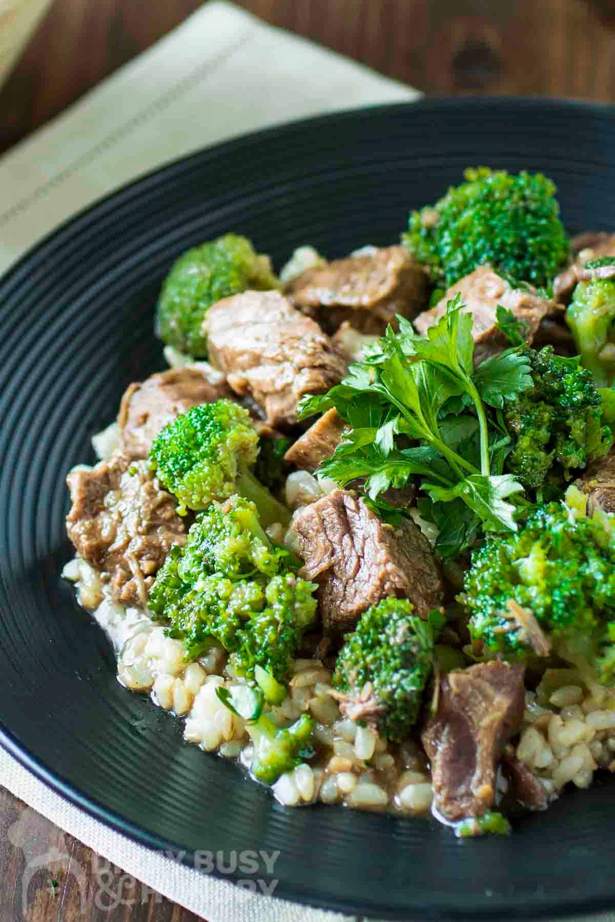 Side shot of beef and broccoli on a bed of rice garnished with fresh parsley on a black plate.