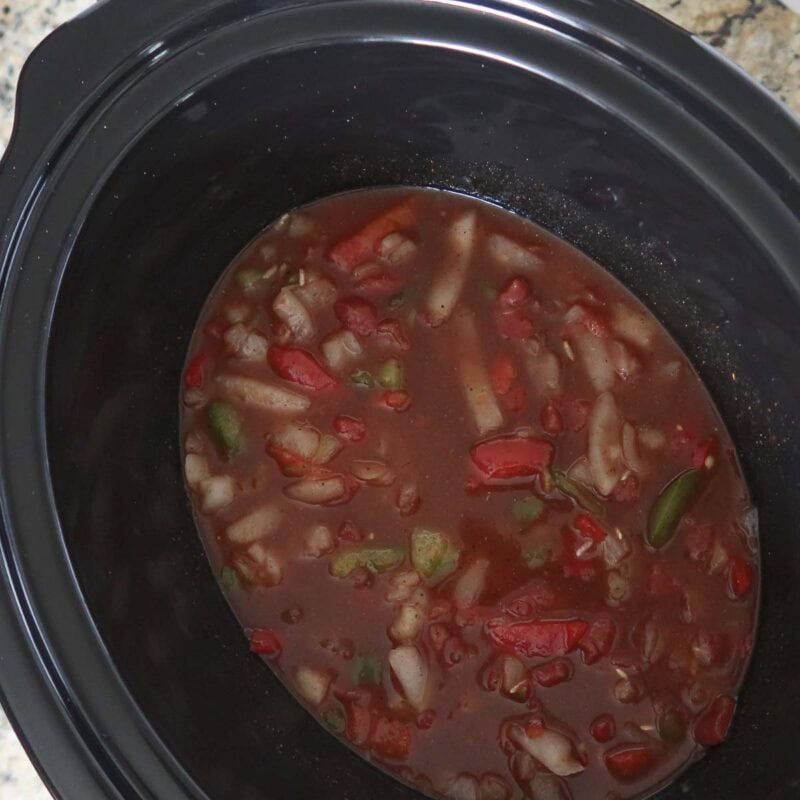 Overhead view of process shot 1 - in the slow cooker, combine the rice, chili powder, cumin, salt, pepper, canned tomatoes, tomato sauce, broth, and frozen pepper and onion strips.