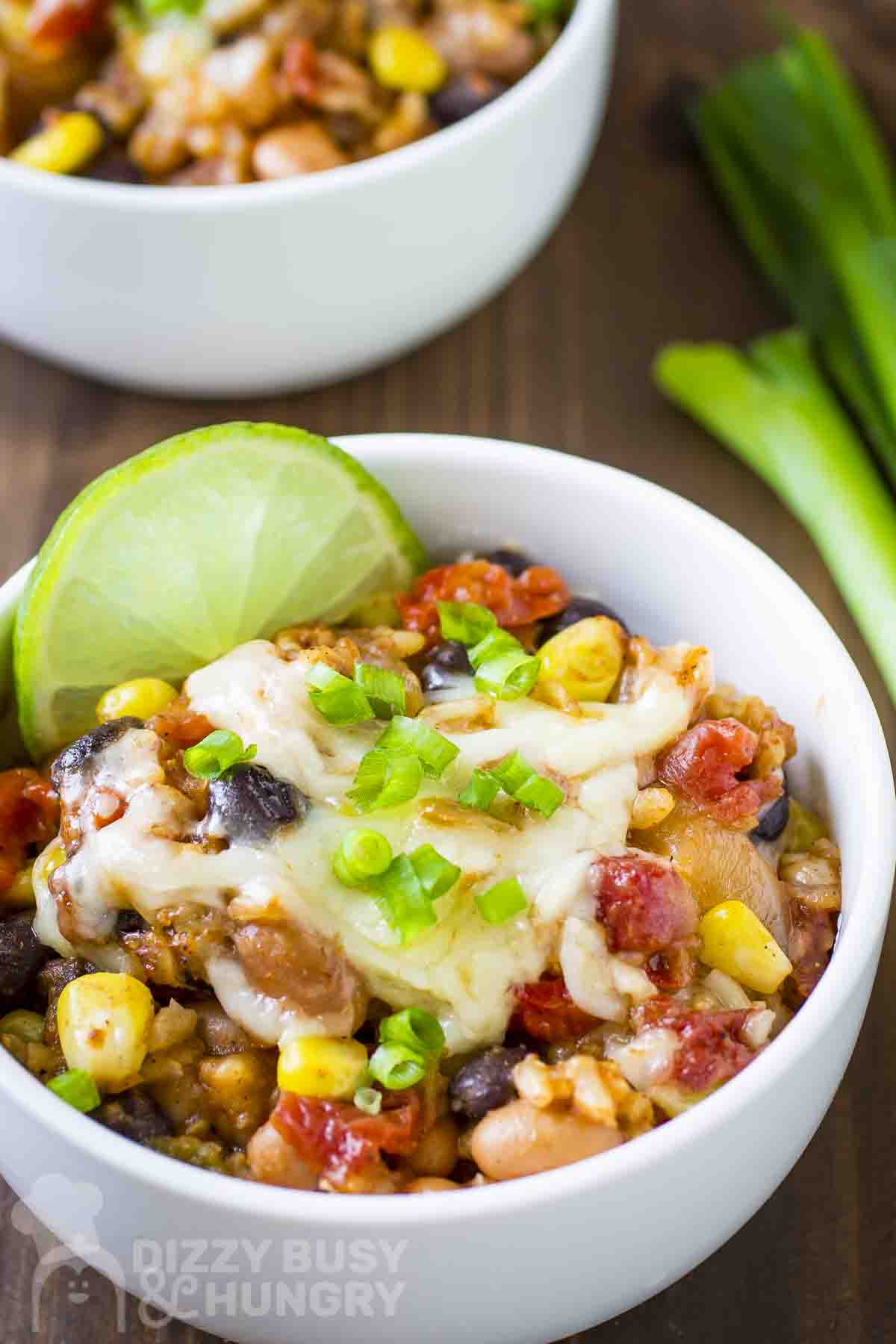 Close up shot of enchilada bake garnished with chopped green onions and a lime wedge in a white bowl on a wood surface.