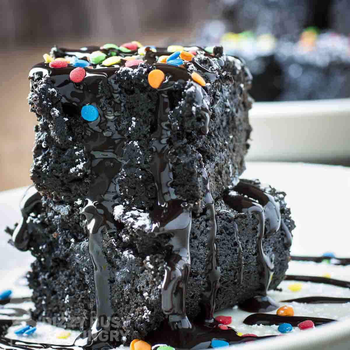 Side shot of two black bean brownies stacked on each other spinkled with powdered sugar, chocolate syrup, and rainbow sprinkles on a white plate.
