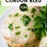 Close up shot of two pieces of chicken cordon bleu garnished with fresh parsley on a white plate with a fork on a green cloth on the side.