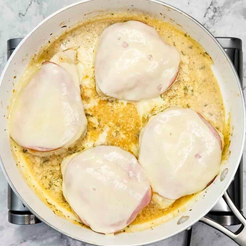 Overhead view of process shot 4 - return the chicken to the skillet and spoon some of the sauce on top of each piece, then top each with a slice of ham and a slice of cheese, then cover the skillet with a lid and cook on low for 3-4 minutes, or until the cheese melts.