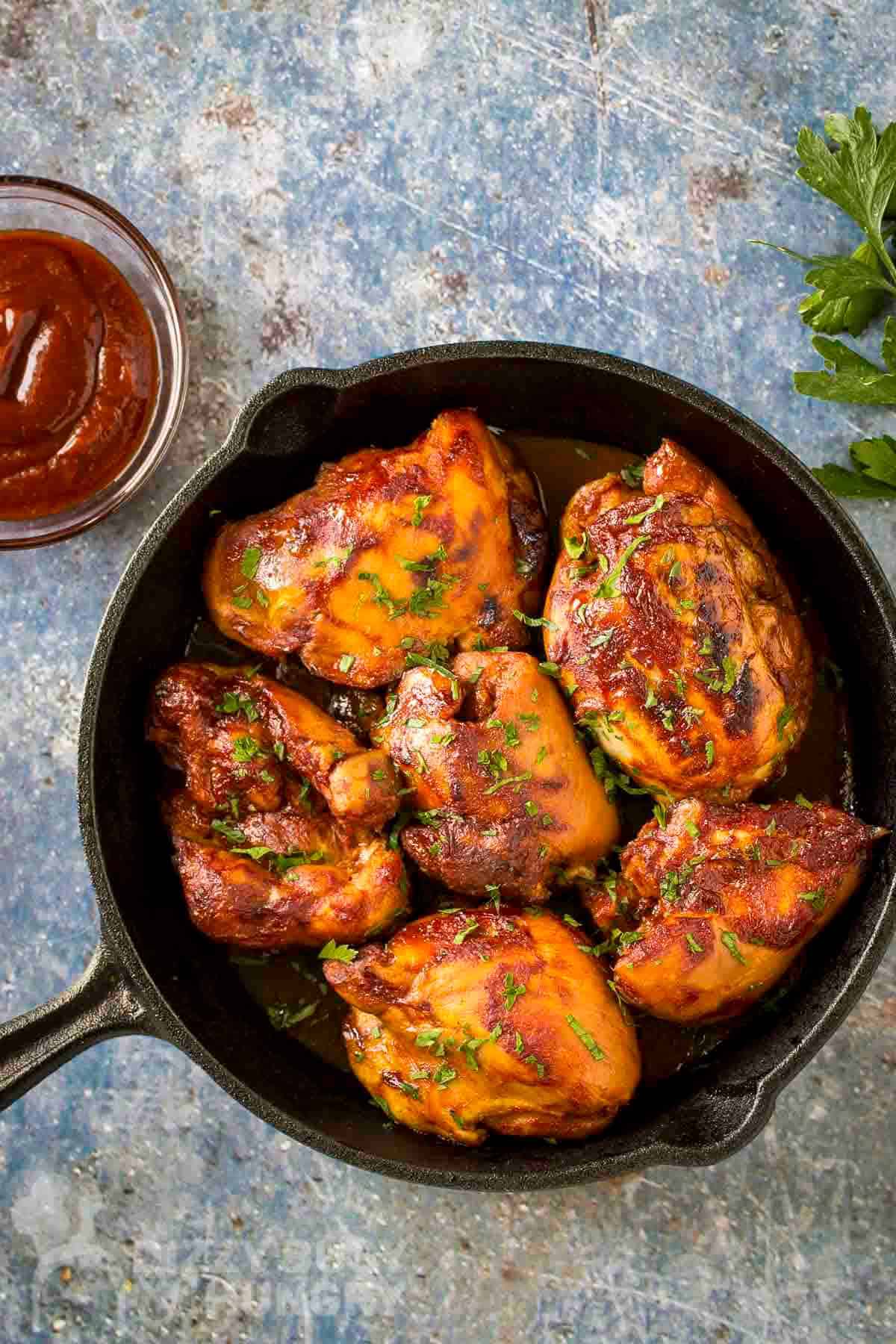 Overhead shot of BBQ chicken thighs in a cast iron skillet garnished with fresh parsley with more fresh parsley sprigs in the background.