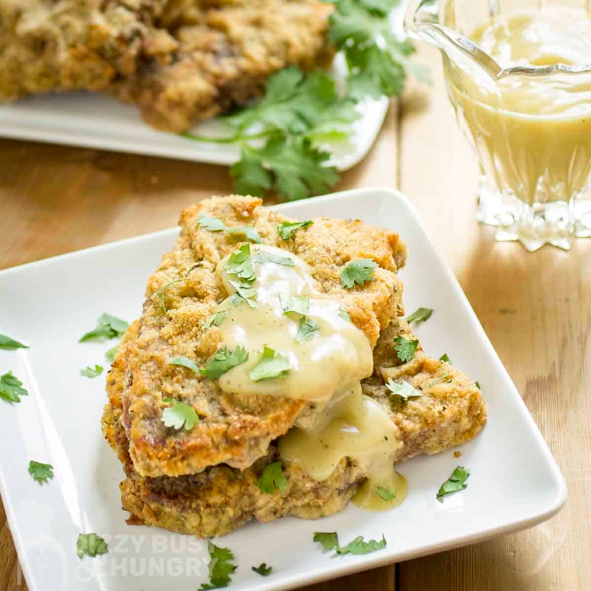 Side shot of chicken fried steak with gravy and fresh parsley on top on a white plate with more gravy in a glass dish on the side.