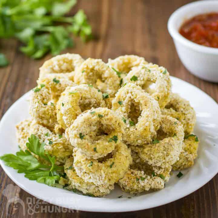 Side view of baked calamari with herbs on a white plate with a bowl of sauce in the background.