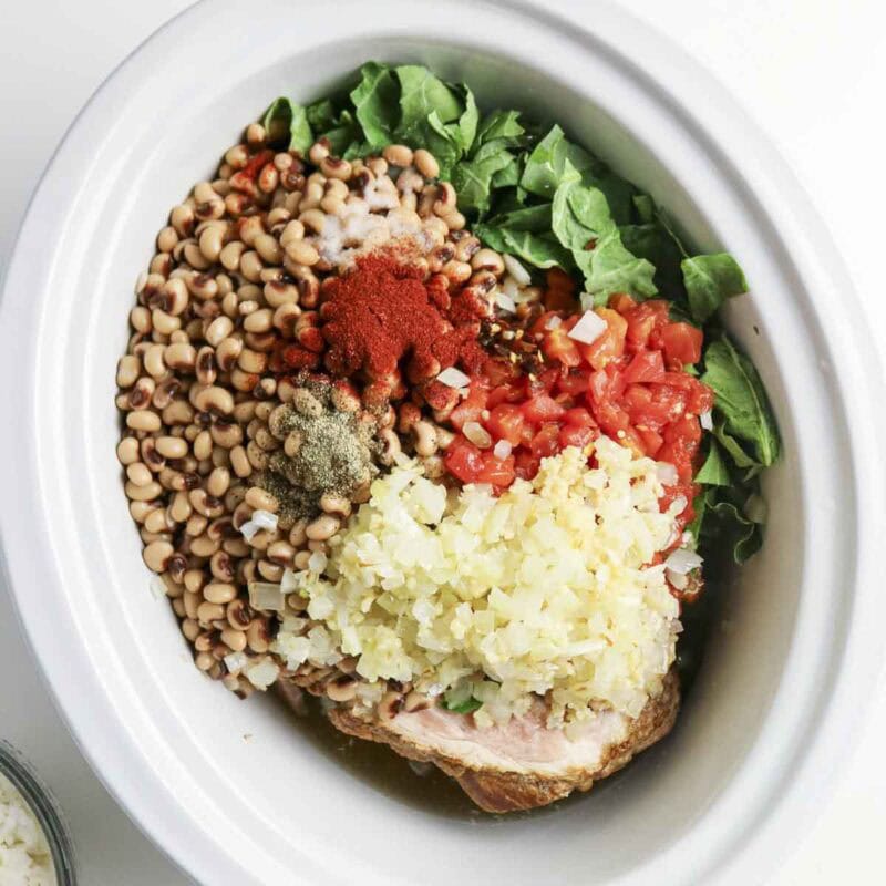 Overhead view of process shot 2 - place the collard greens, black-eyed peas, diced tomatoes, chicken broth, ham bone, smoked paprika, black pepper, salt, and red pepper flakes to the slow cooker. Add the cooked onion and garlic as well. Stir to combine.