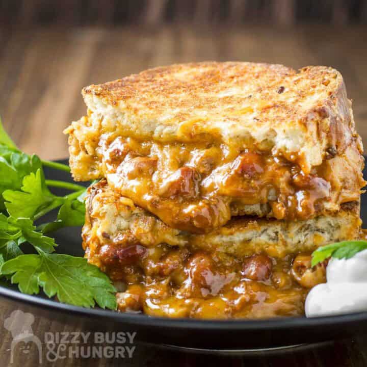 Side shot of two halves of a chili cheese grilled sandwich on a black plate with sour cream and parsley on the sides.