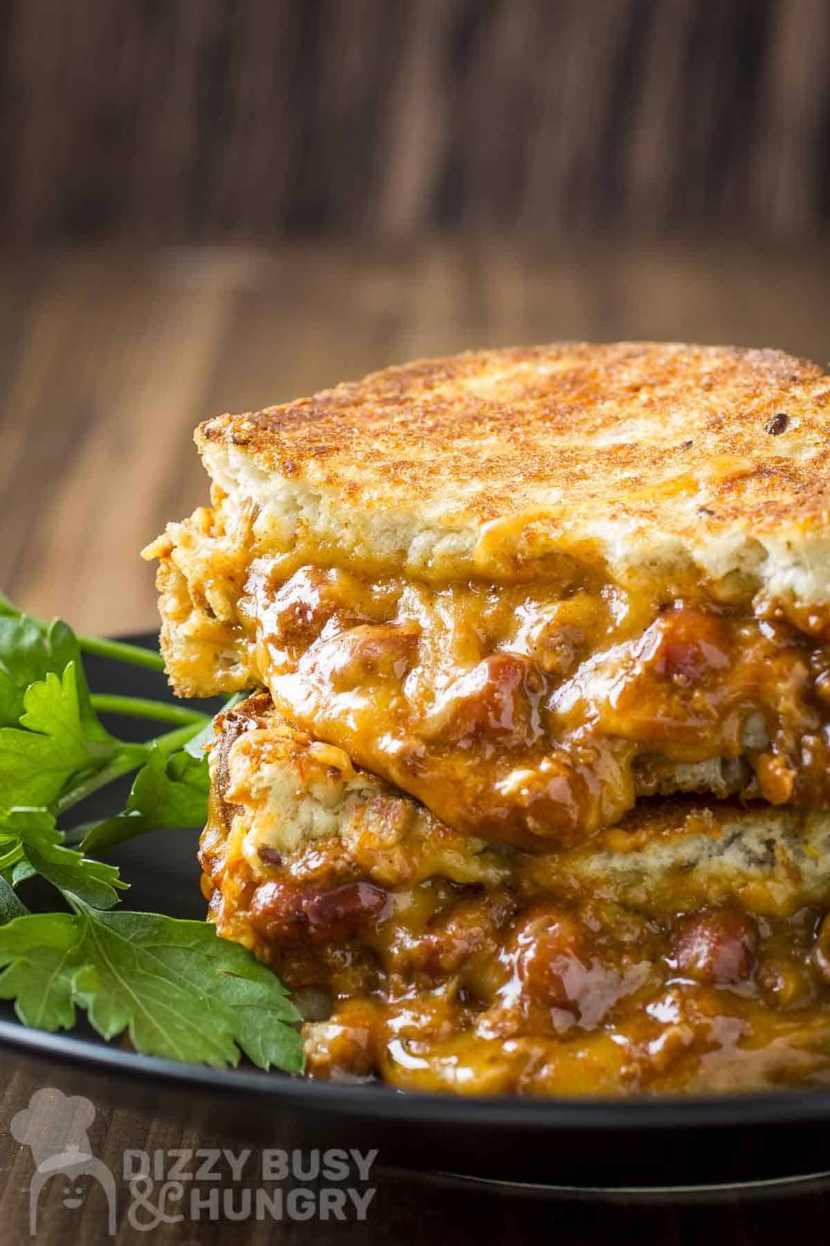 Side shot of two halves of a chili cheese grilled sandwich on a black plate with sour cream and parsley on the sides.
