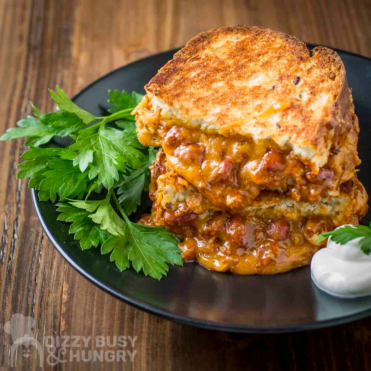 Side shot of two halves of a chili cheese grilled sandwich on a black plate with sour cream and parsley on the sides.