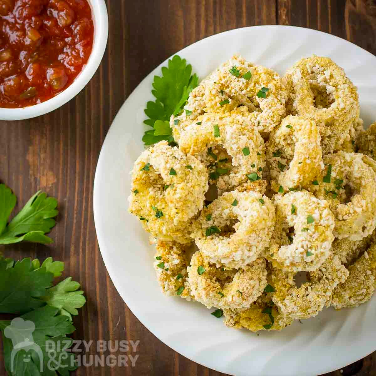 Overhead view of baked calamari with herbs on a white plate with a bowl of sauce in the background.